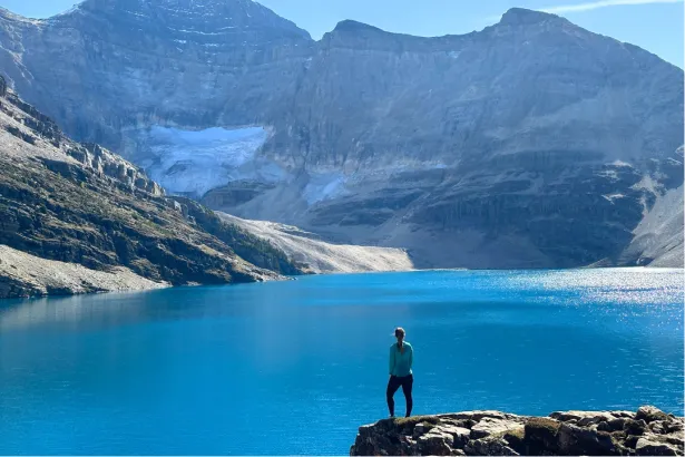 Sunny day at Lake O’Hara with hiker standing on rock overlooking large alpine lake and hazy mountain range in Yoho National Park